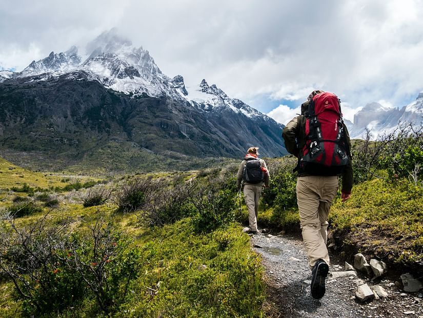Two people walking on a trekking path with greenery around them and a snow-clad mountain in the background